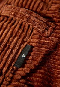 Bruine ribfluweelstof met verticale ribben, voorzien van een textuur oppervlak en een zwart etiket met een logo aan de zijkant.