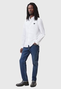 Camisa blanca de botones con un logo de corazón negro, combinada con jeans de mezclilla azul y botas de tobillo negras. Líneas limpias y corte clásico.