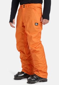 Oranžové lyžiarske nohavice s lesklým povrchom, so zipsovými vreckami, elastickými manžetami a emblémom na boku. Nosené s čiernymi lyžiarskymi čižmami.