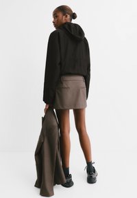 Black hooded jacket, gray mini skirt with pockets, holding a brown blazer, black sneakers, and black socks, viewed from behind.