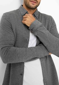 Grau gemusterte Hemd mit Knöpfen, Kragen, langen Ärmeln und dunklen Knöpfen, über einem weißen T-Shirt getragen. Strukturiertes Gewebe.