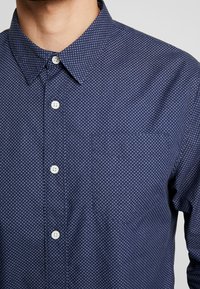 Camicia blu navy con bottoni, caratterizzata da un delicato motivo a pois, colletto aperto e taschino sul petto. Presenta bottoni bianchi e una texture della stoffa liscia.
