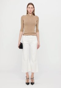 Pull en maille beige avec un col zippé, associé à un pantalon large blanc. Sac à main noir et escarpins complètent la tenue.