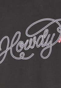 Tejido negro con la palabra "Howdy" bordada en letras blancas en forma de cuerda. El diseño es central y ocupa la mayor parte del espacio.