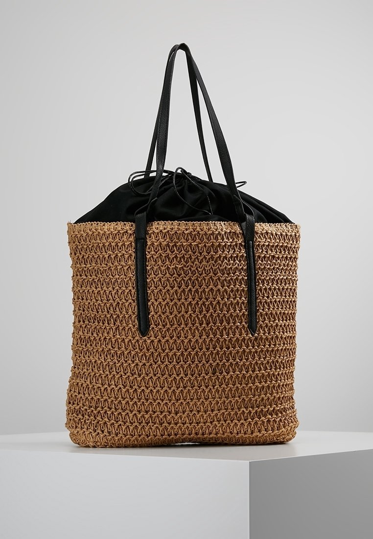 Sac fourre-tout en paille tissée avec une fermeture à cordon noir en tissu et de longues bandoulières noires. Couleur naturelle texturée avec un dessus lisse.