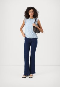 Top de encaje azul claro con mangas de volante, combinado con jeans acampanados azul oscuro y bailarinas beige. Bolso negro sostenido en una mano.