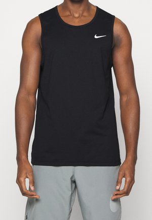 Homme portant un débardeur noir Nike et un short gris clair, debout devant un fond clair uni.