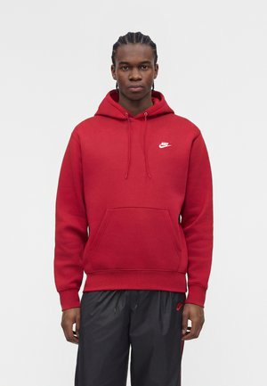 Jeune homme aux cheveux tressés portant un sweat à capuche rouge Nike et un pantalon noir Nike, debout devant un fond blanc uni.
