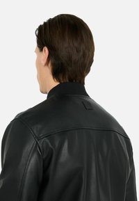 Zwarte leren jas met een gladde textuur, geribde kraag en stiksels op de schouders. Voorzien van een subtiele logopatch op de rug.