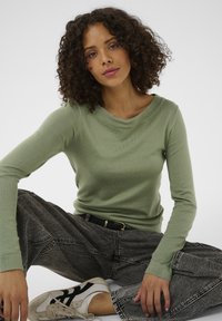 Ženska s kodrastimi lasmi sedi s prekriženimi nogami, oblečena v svetlo zeleno majico z dolgimi rokavi, črni pas, svetlo oprane kavbojke in bele superge.