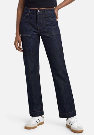 Personne portant un jean bleu foncé à jambes droites avec des poches plaquées devant et des baskets blanches avec des bandes noires.