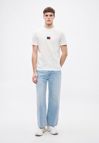 Λευκό βαμβακερό μπλουζάκι με μαύρο και κόκκινο λογότυπο, σε συνδυασμό με τζιν παντελόνι φαρδύ σε ανοιχτό μπλε χρώμα. Το μοντέλο φορά λευκά αθλητικά παπούτσια.