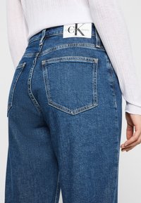 Tamne traperice tamnoplave boje, sa standardnim dizajnom od pet džepova. Imaju Calvin Klein oznaku na pojasu i vidljive detalje šivanja.