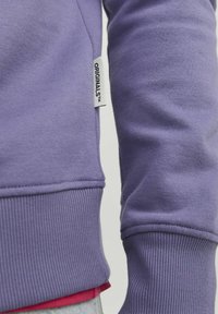 Sudadera púrpura con puños y dobladillo acanalados, que presenta una pequeña etiqueta "ORIGINALS". El material parece suave, con un acabado liso y texturizado.
