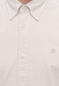 Gros plan sur une chemise à rayures rose clair et blanc avec des boutons, un col boutonné et un petit logo brodé sur la poitrine.