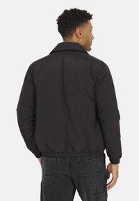 Giubbotto bomber nero con una vestibilità rilassata, caratterizzato da un colletto curvo, polsini e vita elasticizzati, abbinato a pantaloni grigi scuri.