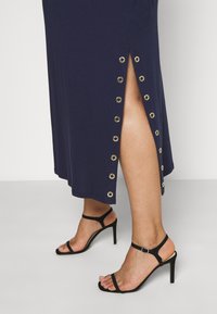 Navy maxi dress with side slit, featuring gold eyelet accents along the hem. Paired with black strappy high-heeled sandals.