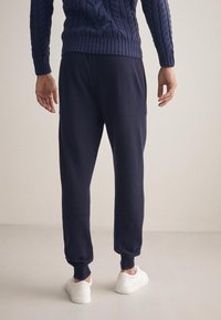 Marinblå stickad tröja med kabelmönster, kombinerad med mörka joggers med elastiska mudd. Modell med vita sneakers, sedd bakifrån.