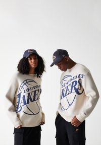 Puloverele tricotate crem au un grafism "Los Angeles Lakers" în nuanță de bleumarin, asortate cu pantaloni cargo negri și șepci bleumarin cu logo-ul Lakers.