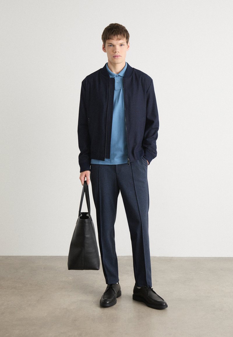 Granatowa kurtka bombowca na jasnoniebieskiej koszulce polo, w połączeniu z ciemnoniebieskimi spodniami i czarnymi butami. Trzyma czarną skórzaną torbę.