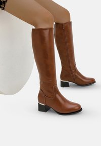 Mujer con botas de cuero marrón hasta la rodilla, con cremalleras laterales y tacones bajos, sentada sobre una superficie blanca y redondeada.