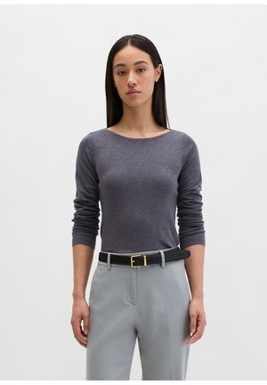 Kobieta z długimi ciemnymi włosami, ubrana w dopasowany szary top z długim rękawem włożony w jasnoszare spodnie z czarnym paskiem.