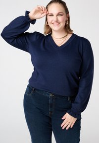 Marineblauwe trui met lange, opgeblazen mouwen en een geschulpte V-hals. Gestructureerde stof en aansluitend ontwerp, gecombineerd met donkerblauwe jeans.