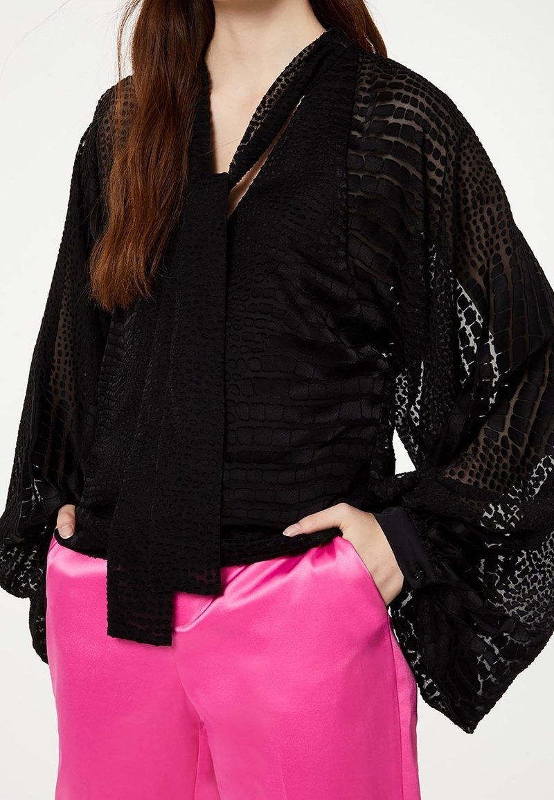 Nainen, jolla on yllä musta teksturoitu pitkähiha blouse, jossa on solmukohta, ja kirkkaan vaaleanpunaiset satiinipöksyt, kädet taskuissa.