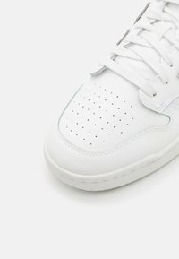 Sneaker bianca in pelle con design perforato, lacci piatti e dettagli di cucitura sottili. Punta arrotondata e suola in gomma morbida.