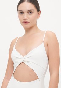 Maillot de bain blanc avec un design à l'avant enroulé, des fines bretelles et un détail découpé à la taille. Tissu texturé avec un motif subtil.