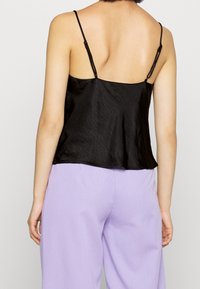 Černý saténový top s tenkými nastavovacími ramínky, který má splývavý střih a mírně nařasený zadní díl. Nosený s světle fialovými kalhotami.