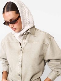 Nő teknősbéka mintás napszemüveget, krémszínű kötött kapucnit és bézs, túlméretezett, gombos inget visel, amelynek az ujjai fel vannak hajtva.