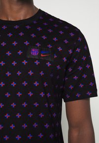 Camiseta negra de manga corta con un patrón de pequeñas cruces rojas y azules. Incluye un parche con los logotipos de Barcelona y Nike.