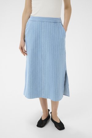 Kvinde iført blå midi-nederdel med lodrette striber og sidesplit, hvid ærmeløs top og sorte net slip-on sko, stående på hvidt gulv.