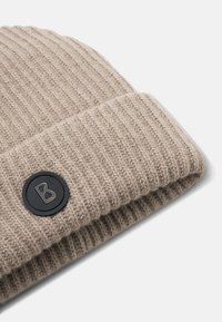 Beige strikket hue med ribbet tekstur, der har en sort cirkulær logo-patch med et bogstav 'B' på den foldede kant.