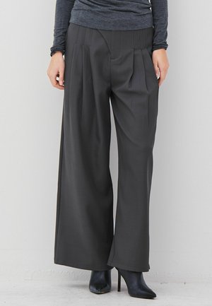 Person mit weit geschnittenen dunkelgrauen Hosen, schwarzen hohen Stiefeletten und einem enganliegenden grauen Langarmoberteil vor einem weißen Hintergrund.