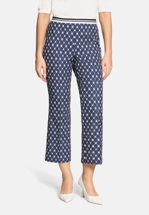 Kobieta ubrana w granatowe, krótkie spodnie z białym geometrycznym wzorem, zestawione z białymi szpilkami w noskiem i białą fakturowaną bluzką.