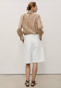 Une femme se tient de dos à la caméra, portant un chemisier beige et un short en jean blanc, associé à des sandales noires à lanières.