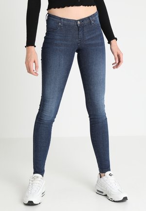 Nainen pukeutunut tummansinisiin skinny-farkkuihin, valkoisiin lenkkareihin ja mustaan lyhyt pusero, seisoo valkoista taustaa vasten.