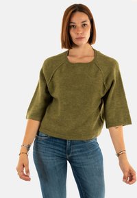Pull court olive, à manches courtes, avec un large col et une coupe décontractée, fabriqué à partir d'un matériau tricoté doux et texturé. Associé à un jean bleu.