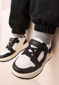 Child wearing black and white sneakers with Velcro straps and gray socks, standing against a light-colored wall.