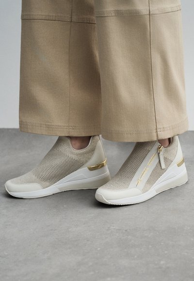 Baskets slip-on beiges avec un dessus en tricot texturé, semelle blanche, accents en matériel doré et un détail de fermeture éclair sur le côté.