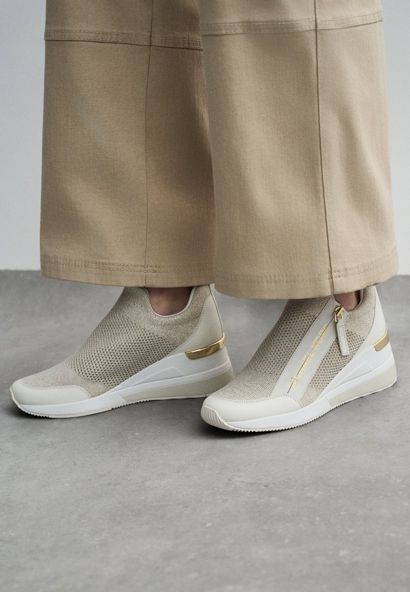 Beige Slipper-Sneaker mit strukturiertem Strick-Obermaterial, weißer Sohle, goldenen Hardware-Akzenten und einem Reißverschlussdetail an der Seite.