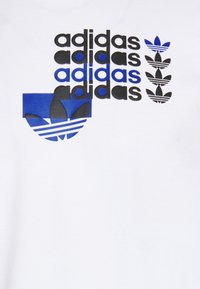 Bílá tričko se spoustou černých a modrých nápisů "adidas" a logem adidas s třemi pruhy na spodní části.