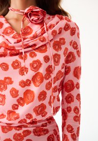 Top floreale a maniche lunghe in tessuto rosa con motivi di rose rosse, caratterizzato da un collo a scialle e un dettaglio di rosa abbinato attaccato al colletto.
