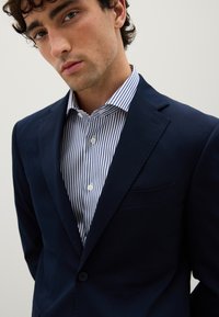 Blazer blu navy con un design strutturato, chiusura a un bottone e una tasca. Indossato sopra una camicia bianca e blu a righe con dettagli sui bottoni.