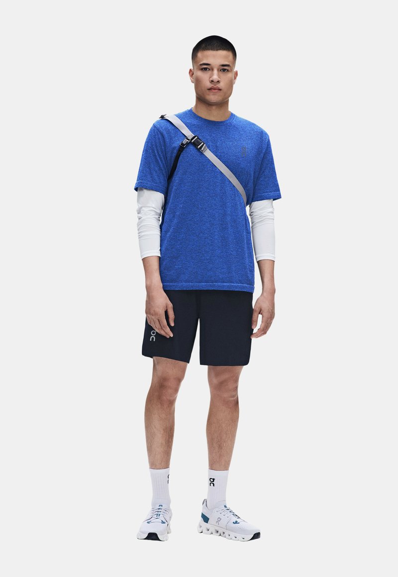 Homme debout portant une chemise bleue à manches courtes par-dessus des manches longues blanches, un short noir, des chaussettes blanches, des chaussures de sport blanches et une bandoulière grise.