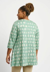 Tunika verde menta con maniche a tre quarti, caratterizzata da un motivo di quadrati bianchi e beige simili a pennellate. Tessuto morbido e leggero.