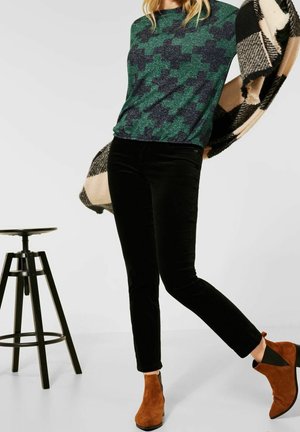 Femme portant un pull à motifs verts et marine, un pantalon noir, des bottines châtaigne, et tenant une écharpe à carreaux beige et noir, se tenant près d'un tabouret en bois.