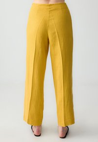 Gele linnen broek met een hoge taille, wijde pijpen en voorplooien. Gladde textuur en minimale naden, rustend boven zwarte sandalen.
